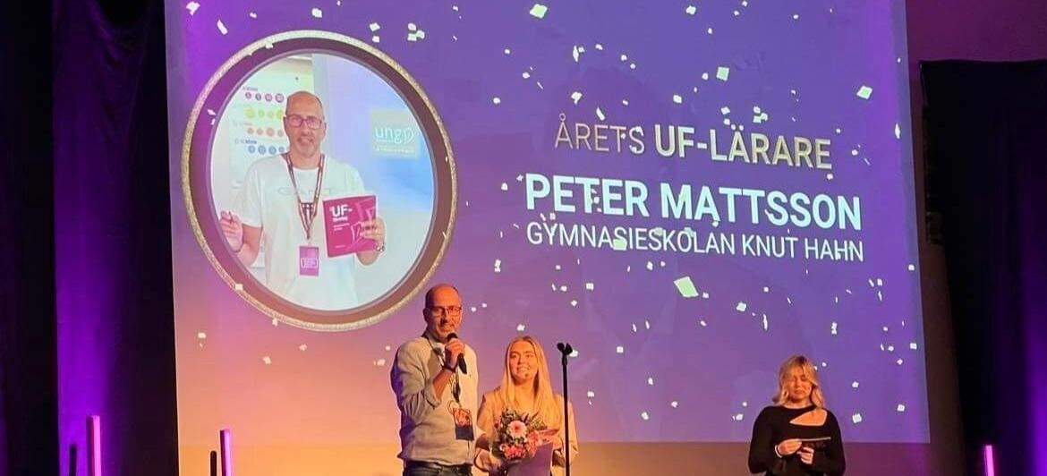 Tre personer står på en scen i lila ljus. Bakom en projektion med texten Årets UF-lärare Peter Mattsson Gymnasieskola Knut Hahn.