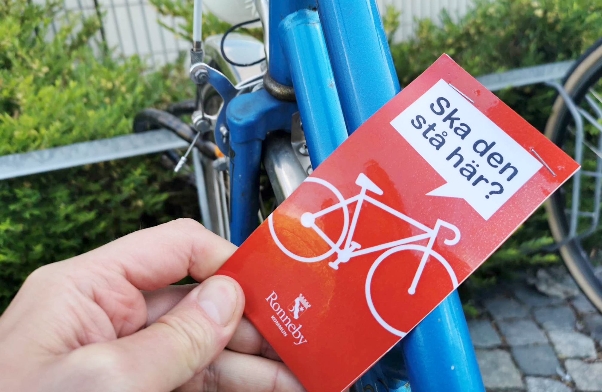 Felparkerad cykel uppmärkt med röd lapp, med budskap/fråga "Ska den stå här?", avsändare Ronneby kommun.